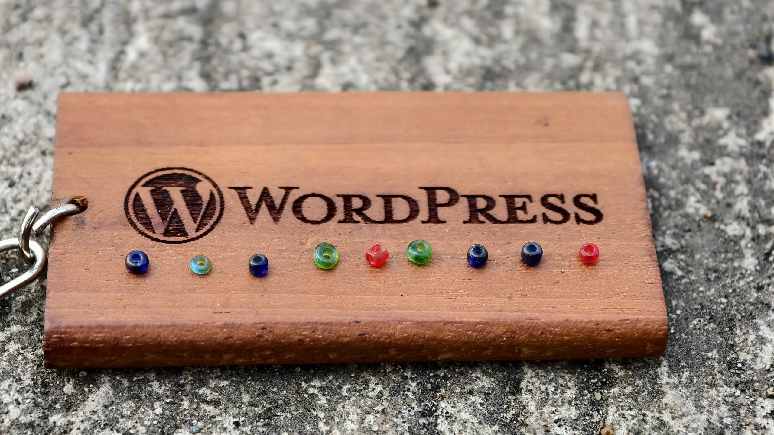 A wooden block with the WordPress logo rests on concrete, with small colorful glass beads lined up in front, shown in